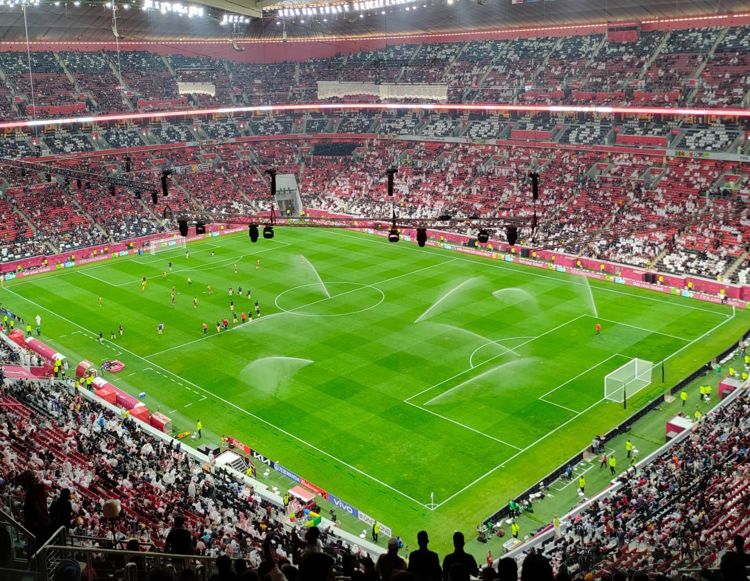 Inside Al Bayt Stadium in Al Khor: red-and-white tiered stands around the pitch as sprinklers water the grass before kick-off